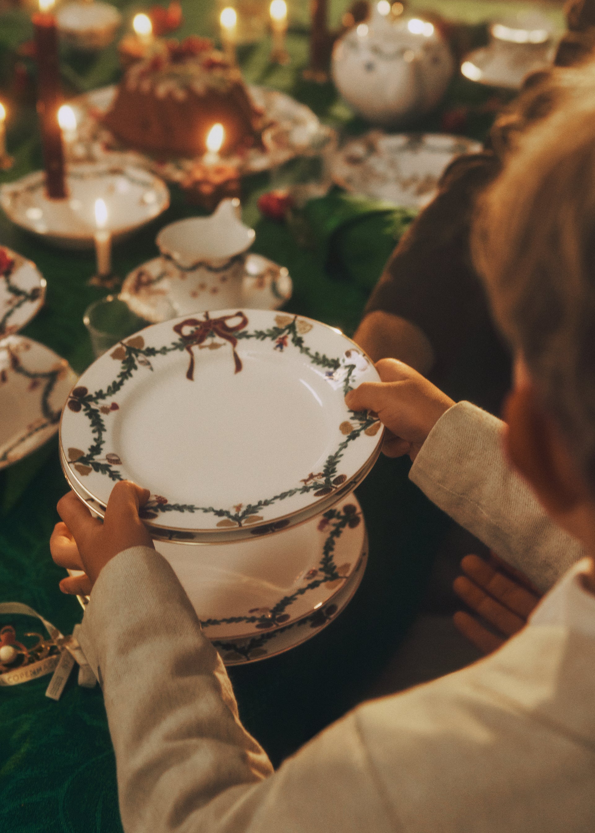 Person holding a stack of Star Fluted Christmas plates. In the background we can see a beautiful set table with Star Fluted Christmas porcelain and illuminated by candle lights.