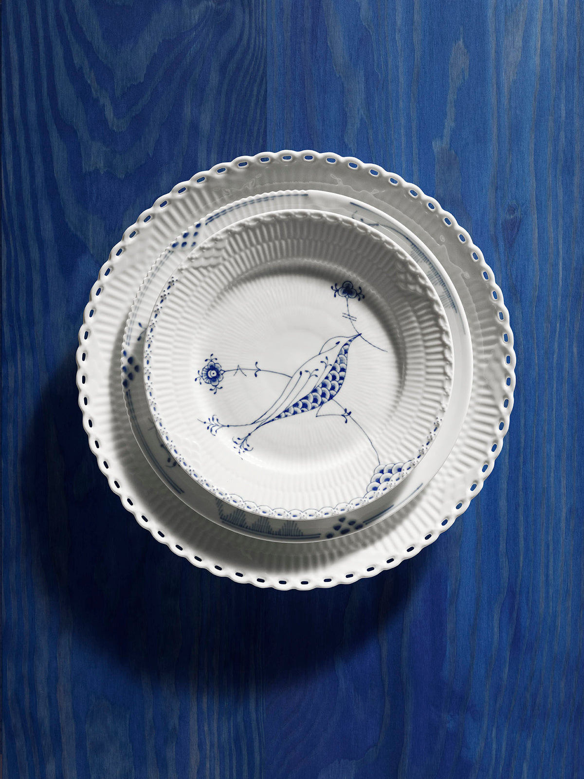 A stack of porcelain plates from Royal Copenhagen including a White Fluted Full Lace plate, A Blue Elements Plate and a Royal Creatures plate on a blue painted wooden table.